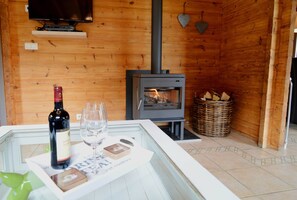 A cozy wooden room with a fireplace, a television mounted on the wall, and a table with a bottle of wine, a glass, and a menu. - Chalet in Ardennes With Sauna & Garden (Waimes)