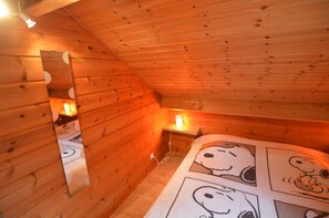 A wooden cabin room with a sloped ceiling, a bed with Snoopy-themed bedding, and a nightstand with a lamp.