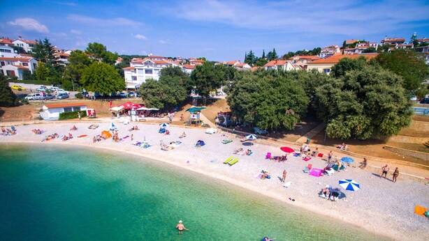 A beach with clear turquoise water, people sunbathing, and a row of buildings in the background. - Comfortable Apartment Near the sea (Medulin)
