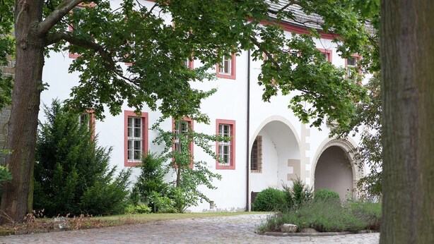A cobblestone pathway leading to a white building with red window frames and arched doorways.