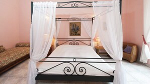 A four-poster bed with white linens, a small bedside table with a lamp, and a wicker chair in a pink-walled room.