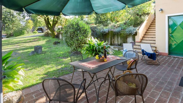 Un patio al aire libre con una mesa y sillas bajo un paraguas grande, rodeado de follaje y una pared de piedra.