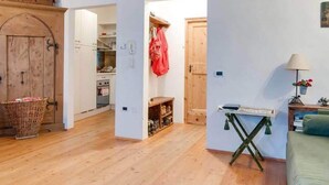 Quarto aconchegante com piso de madeira, sofá verde, uma mesa pequena e um espaço de cozinha com armários de madeira e um casaco vermelho pendurado.