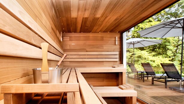 A sauna with wooden benches and a metal bucket, a glass door leading to an outdoor area with chairs and umbrellas. - Opulent Holiday Home in Spa with Sauna, Hot Tub, Playroom (Spa)