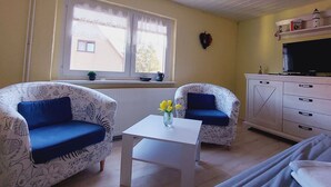 A room with two patterned armchairs, a small white table with a vase of flowers, a television on a cabinet, and a window with a view of a building outside. - Apartment in Kropelin With Terrace (Kroepelin)