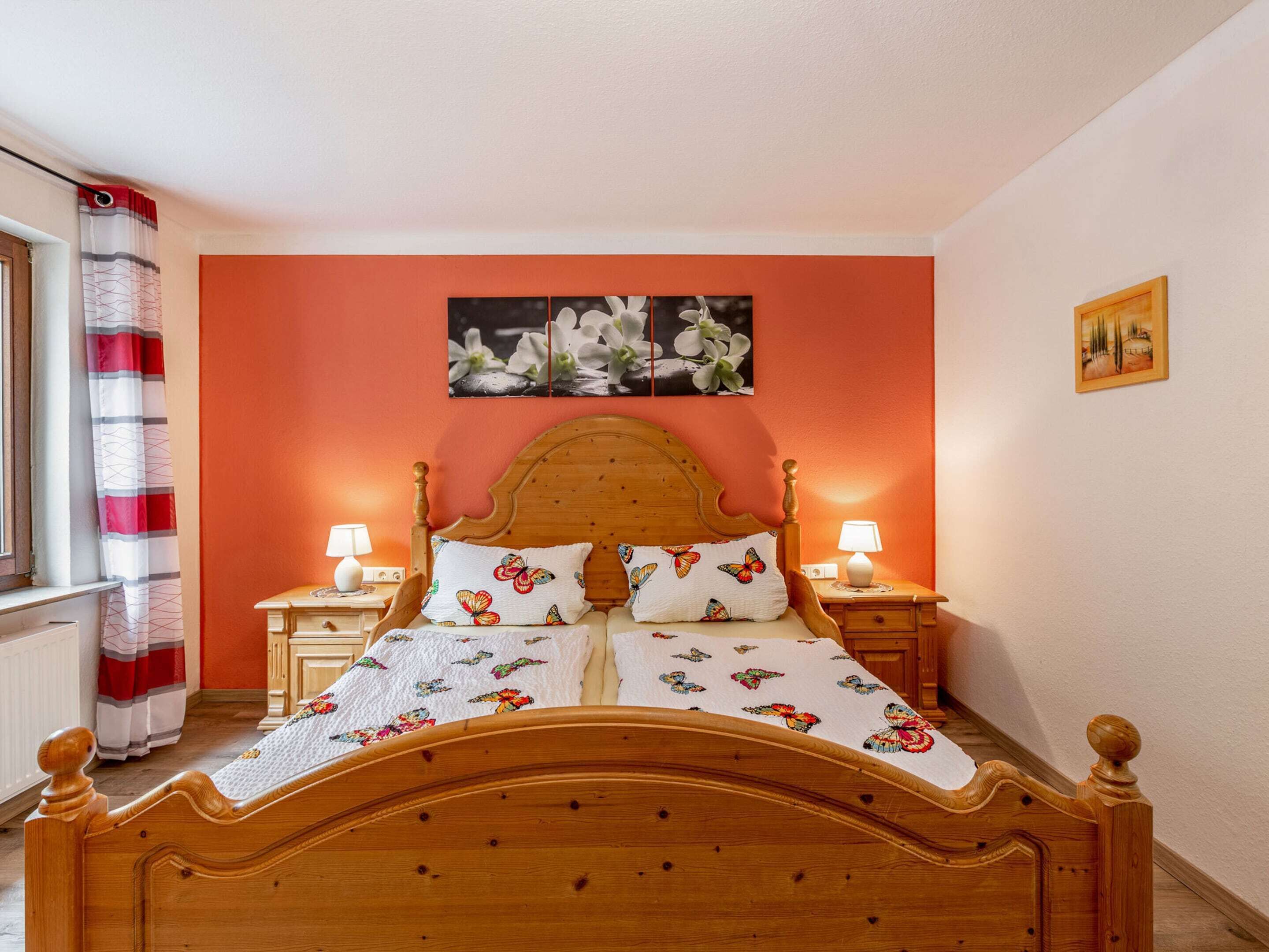 Ein Schlafzimmer mit einem hölzernen Bett, zwei Nachttischen mit Lampen, einem Fenster mit gestreiftem Vorhang und einem Gemälde an der Wand.