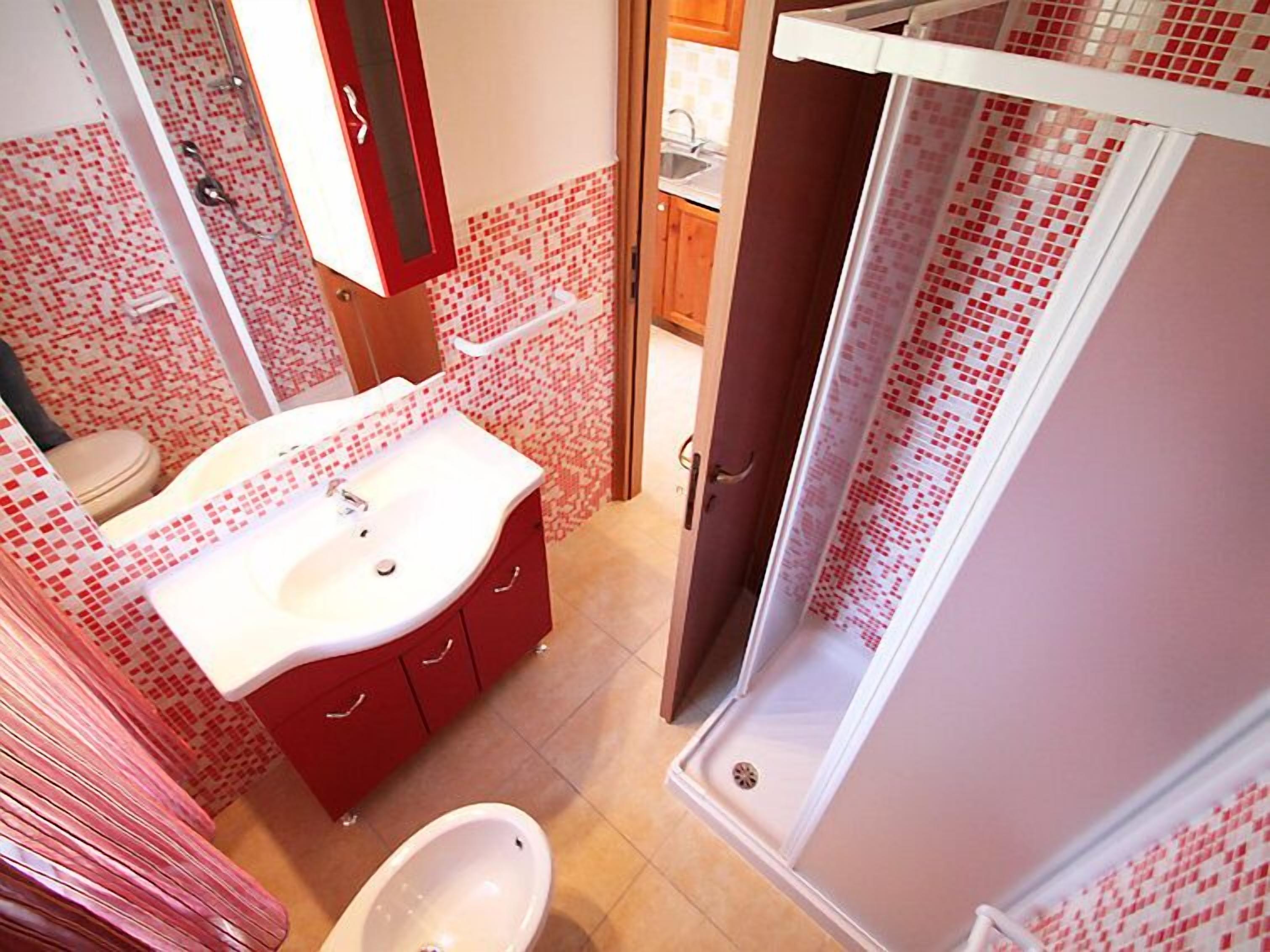 A bathroom with a white sink, red cabinets, a toilet, and a shower with red mosaic tiles.