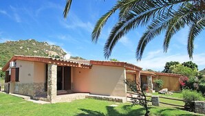 Una casa de un piso con base de piedra y techo de tejas, rodeada de un césped cuidado y palmeras.