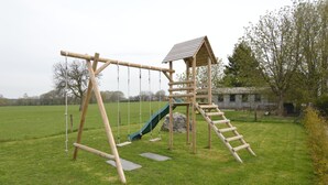 Un ensemble de jeux d’extérieur en bois, comprenant une balançoire et un petit toboggan, installé dans un champ d’herbe.