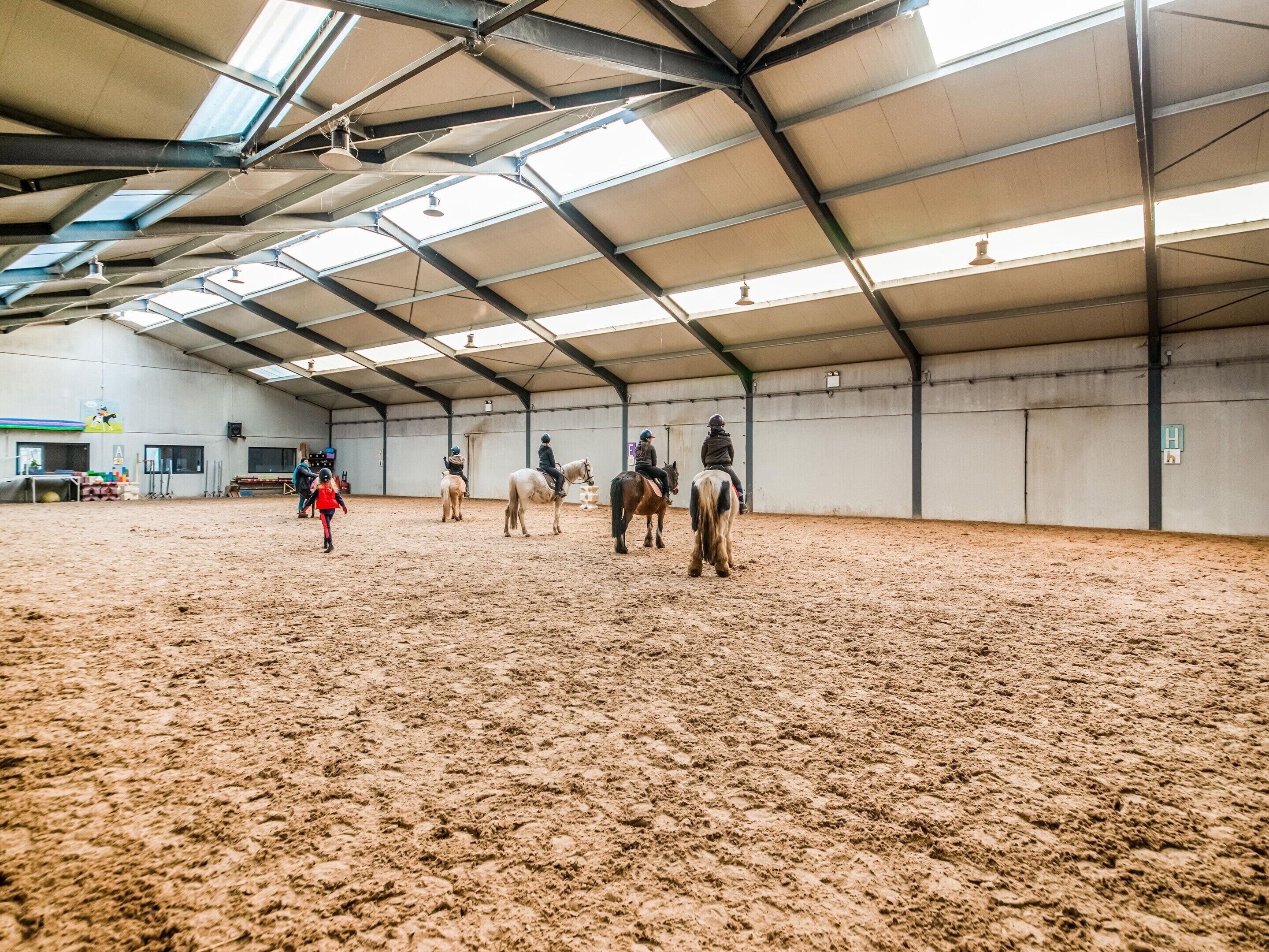 Une carrière d’équitation intérieure dotée d’un sol sablonneux, de poutres métalliques et d’un plafond haut avec des lucarnes.
