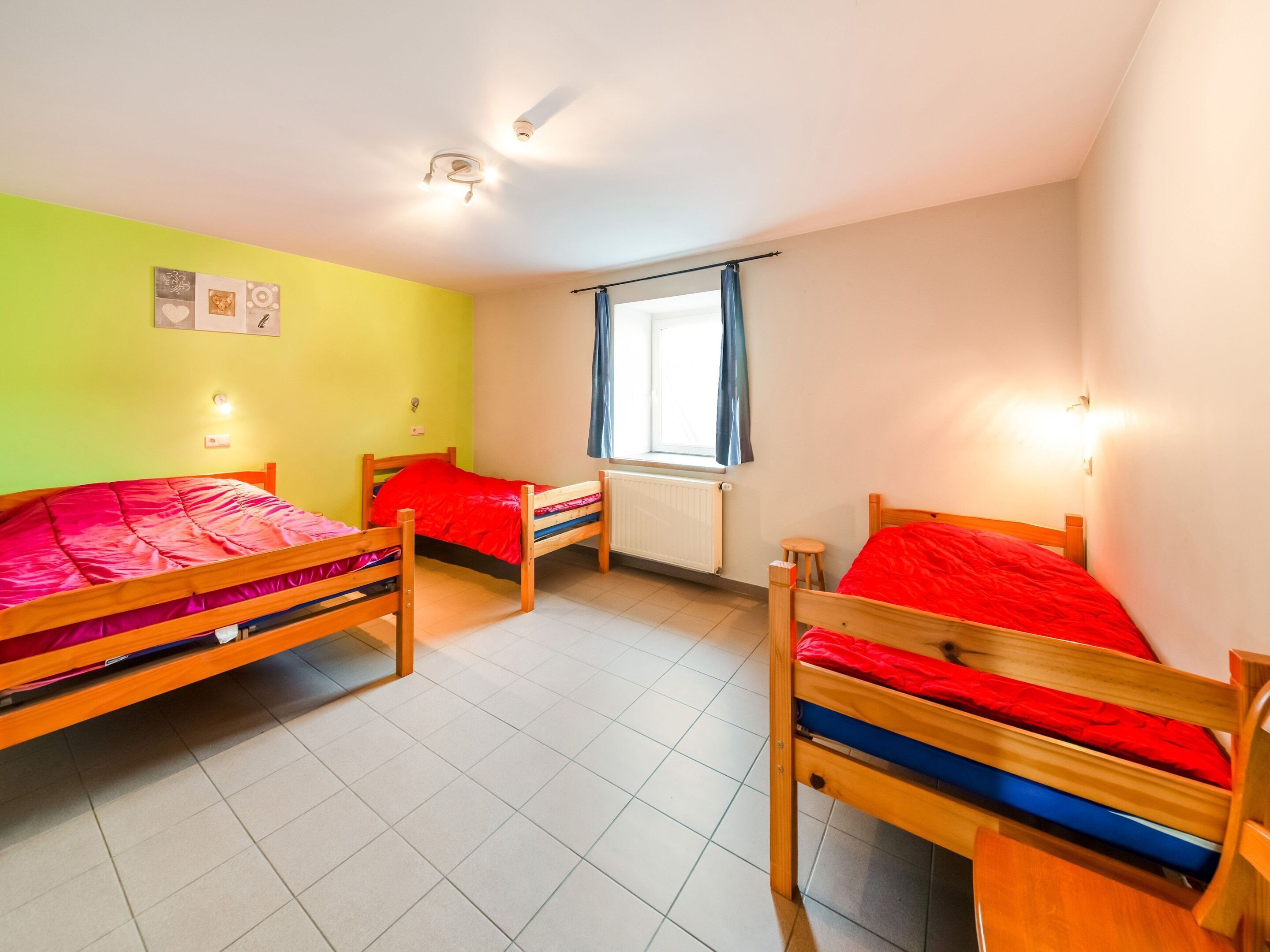 Quarto com três camas de madeira, uma janela com cortinas, um radiador e um candeeiro no teto.