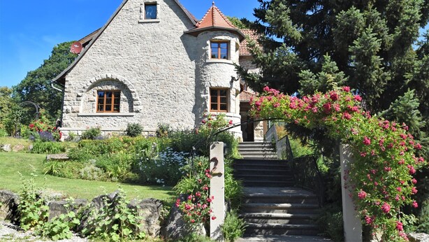 A stone house with a steep roof, a small balcony, and a garden with pink flowers.