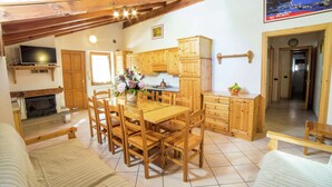 Una cocina acogedora y bien iluminada con armarios de madera, una mesa de comedor y sillas. Un televisor está montado sobre la chimenea y hay un cuadro en la pared.