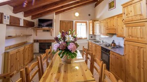 Una cocina con armarios de madera, una mesa de comedor con sillas, un florero con flores y un televisor en un estante.
