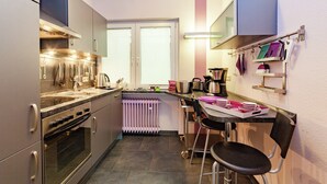 A compact kitchen with a built-in oven, a kettle, and a coffee maker. There is a window with blinds, a radiator, and a chair near the window. - Dog-friendly Holiday Home in Hesse With Garden (Bad Zwesten)