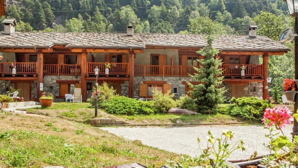 A rustic chalet with a stone facade, wooden balconies, and a garden area with potted plants and a large tree.