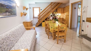 A cozy living room with a sofa, a dining table with chairs, a wooden staircase, and a kitchen area with wooden cabinets.