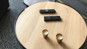Una sala moderna con un sofá gris, un cojín estampado, una mesa de centro redonda de madera, dos controles remotos y dos velas.