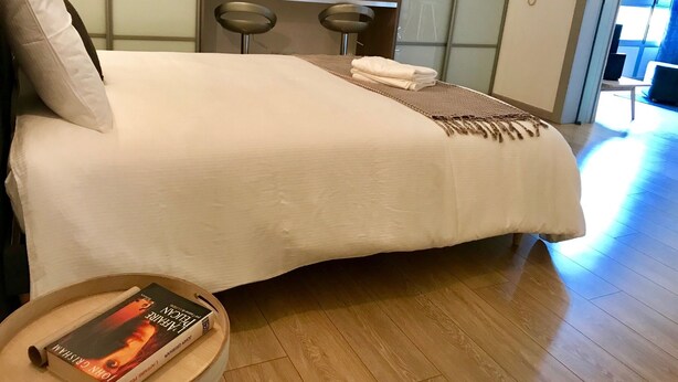 Habitación de hotel moderna con una cama grande, dos mesitas de noche, una pequeña mesa redonda con un libro, y vista al exterior a través de una puerta de vidrio.