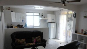 A living room with a black leather sofa and a white coffee table, a kitchen with white cabinets and a refrigerator, and a ceiling fan. - Charming T2 '' Le Colibri '' garden floor with flowers and all comforts in Ste Anne Wifi (STE ANNE)