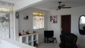 A living room with a black leather chair, a TV, a ceiling fan, and a window with decorative items. - Charming T2 '' Le Colibri '' garden floor with flowers and all comforts in Ste Anne Wifi (STE ANNE)