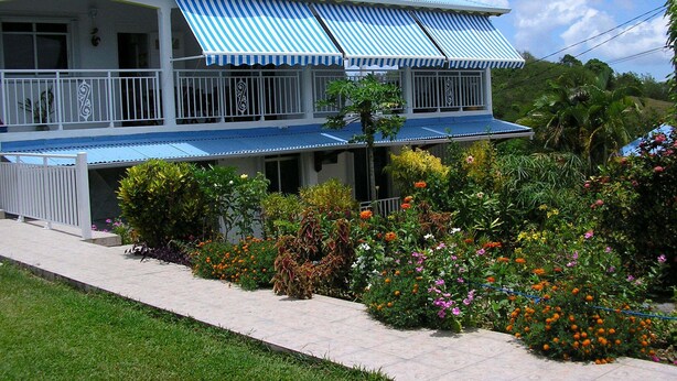 A two-story house with a blue roof and striped awnings, surrounded by a well-maintained garden with various plants and flowers. - Charming T2 '' Le Colibri '' garden floor with flowers and all comforts in Ste Anne Wifi (STE ANNE)
