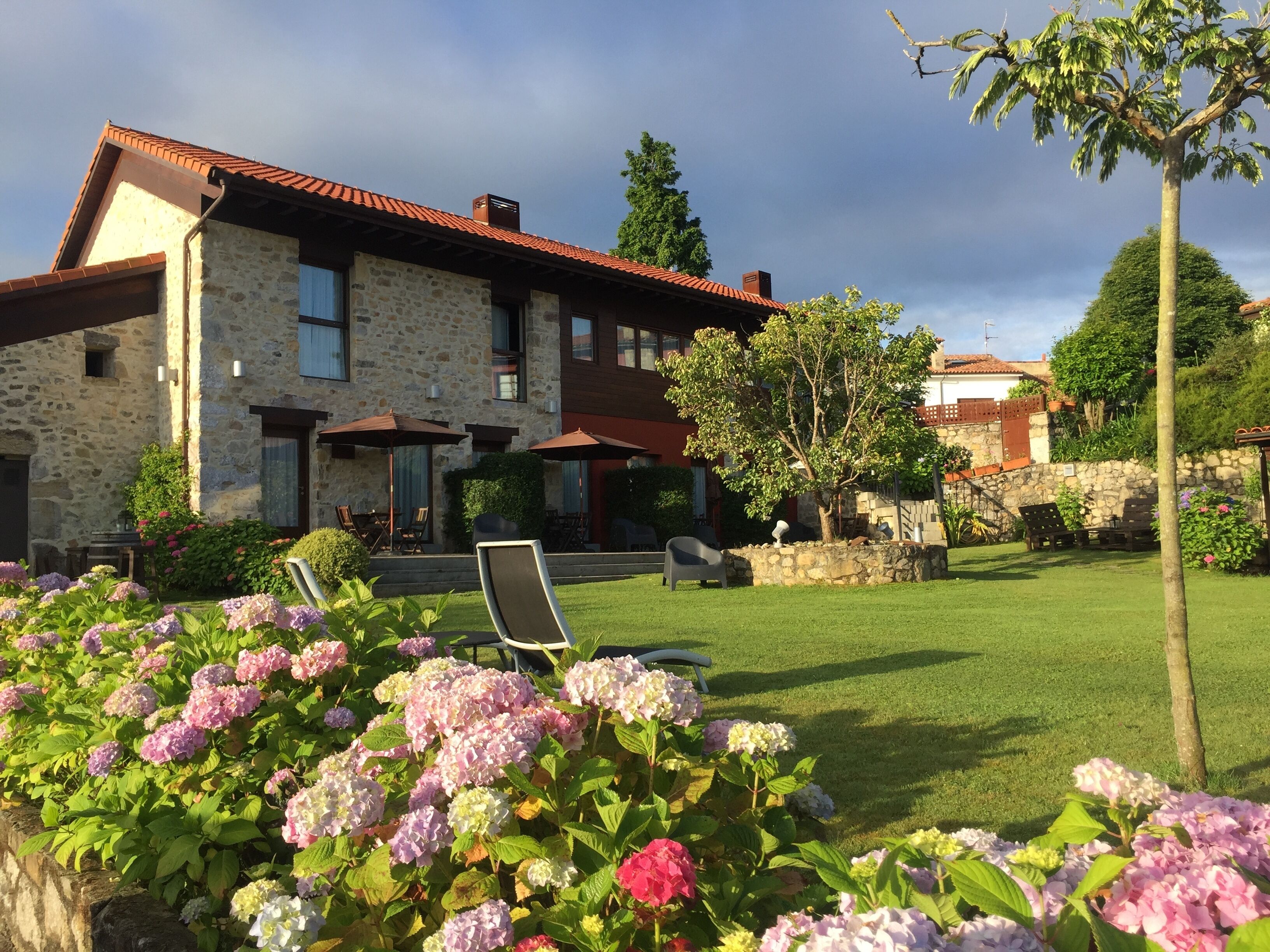 A stone house with a red-tiled roof, a grassy lawn, and a garden with hydrangea flowers.