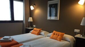 Habitación de hotel con dos camas, cada una con ropa blanca y almohadas naranjas. Hay dos mesitas de noche con lámparas, una ventana con cortinas y un cuadro en la pared.