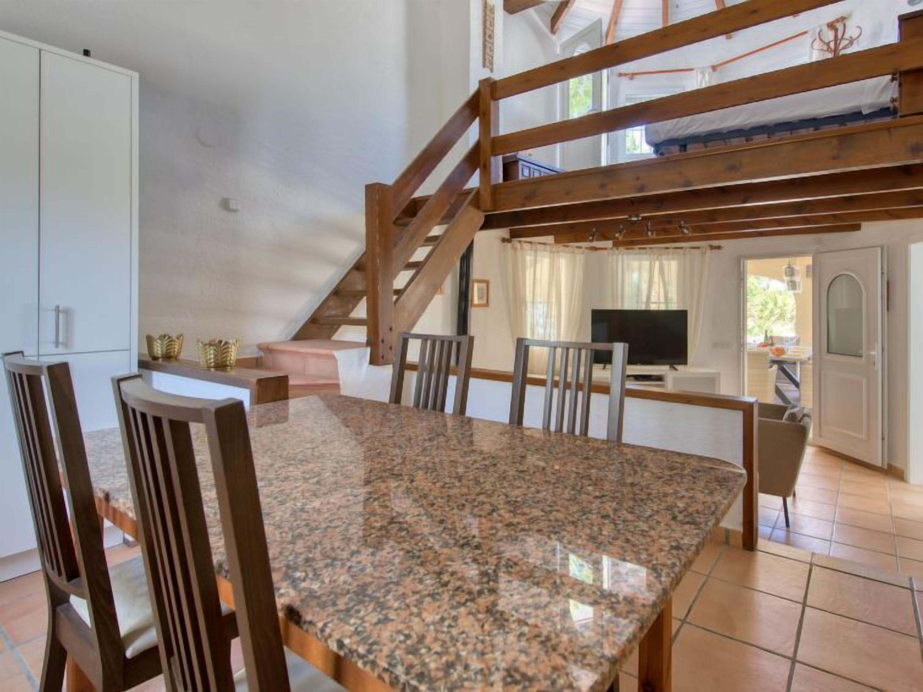 Un'area pranzo con tavolo in granito e sedie in legno, una cucina con mobili bianchi e una scala che conduce a un piano superiore.