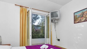 Una habitación de hotel con cama, ventana con cortinas, un cuadro en la pared y un balcón con vistas a árboles y un edificio.