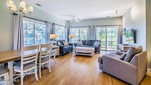 A spacious living room with a wooden dining table, two sofas, and a TV mounted on the wall. - Golf Cart, River and Marsh Views, Oceanfront Community, Beautiful Sunsets (St Helena Island)