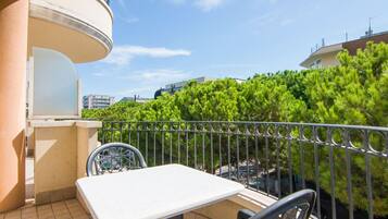 A balcony with a white table and chairs, surrounded by a metal railing, overlooking a green tree and buildings.