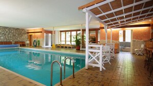 Une piscine intérieure avec un mur en pierre, une terrasse en bois et un espace de détente couvert, équipé de tables et de chaises.