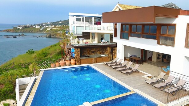 Una casa moderna con una gran piscina, una terraza con sillones de descanso y vista al mar.