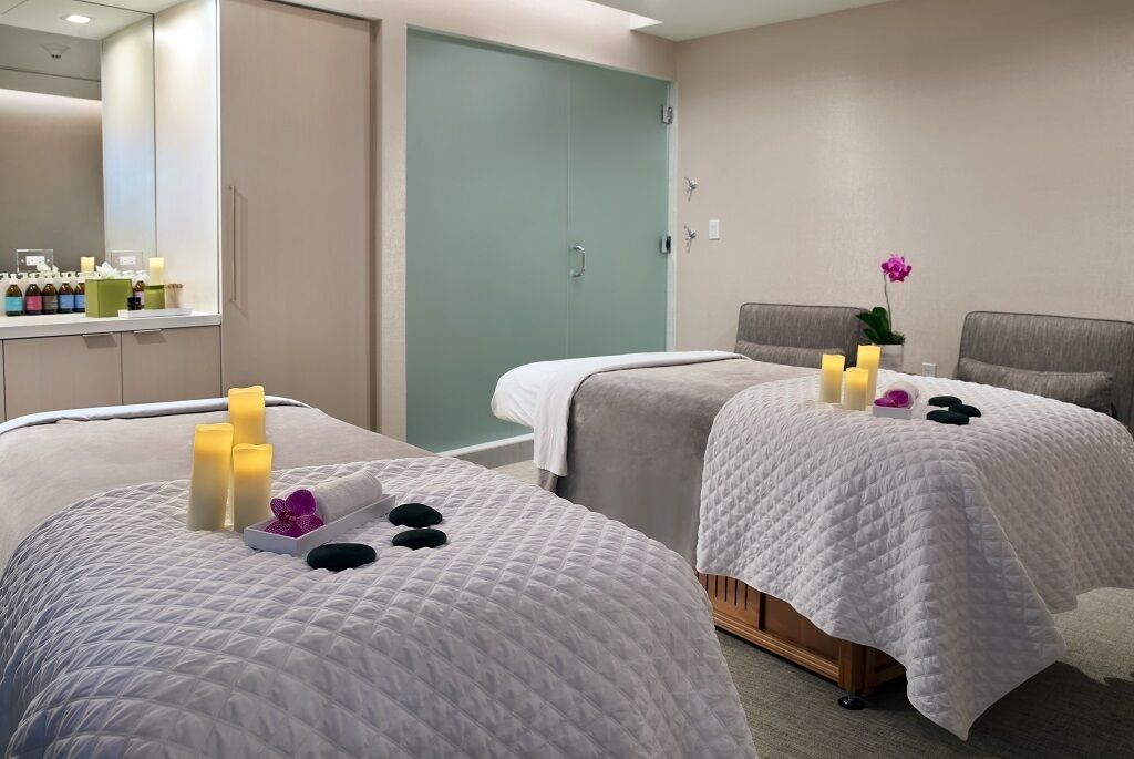 A spa room with two beds, each with a white quilted bedspread, candles, and a small tray with a purple flower.