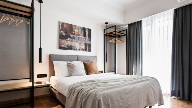 A modern bedroom with a bed, a hanging light, a painting, and a window with curtains. - Modern Minimalist 3BD Apartment in Hilton District (Athens)