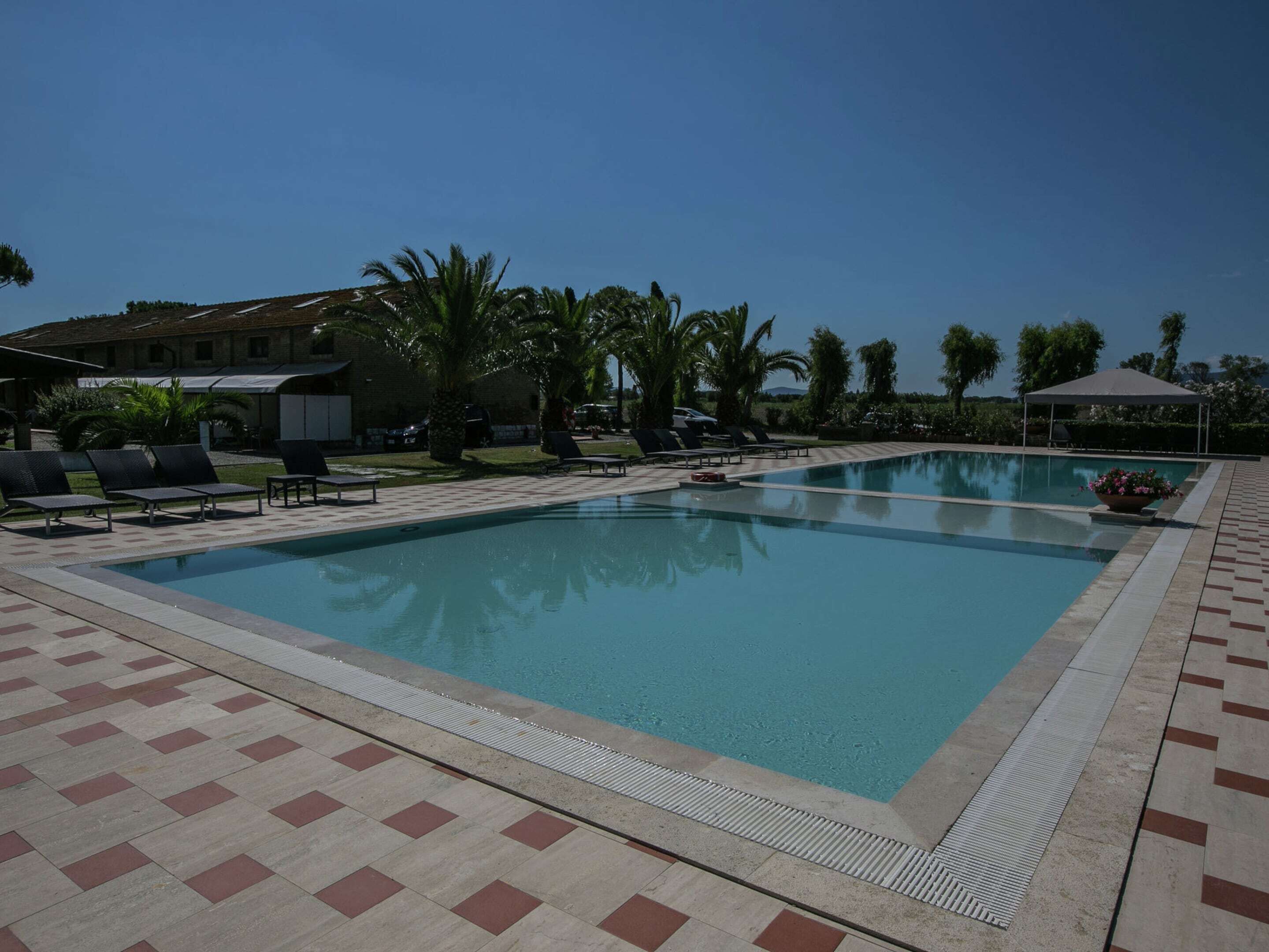 A swimming pool with lounge chairs and a covered area, surrounded by palm trees and a building in the background.