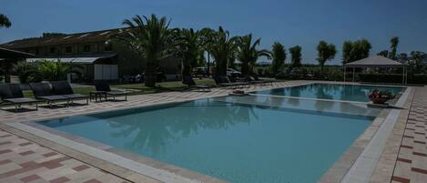 A swimming pool with lounge chairs and a covered area, surrounded by palm trees and a building in the background.