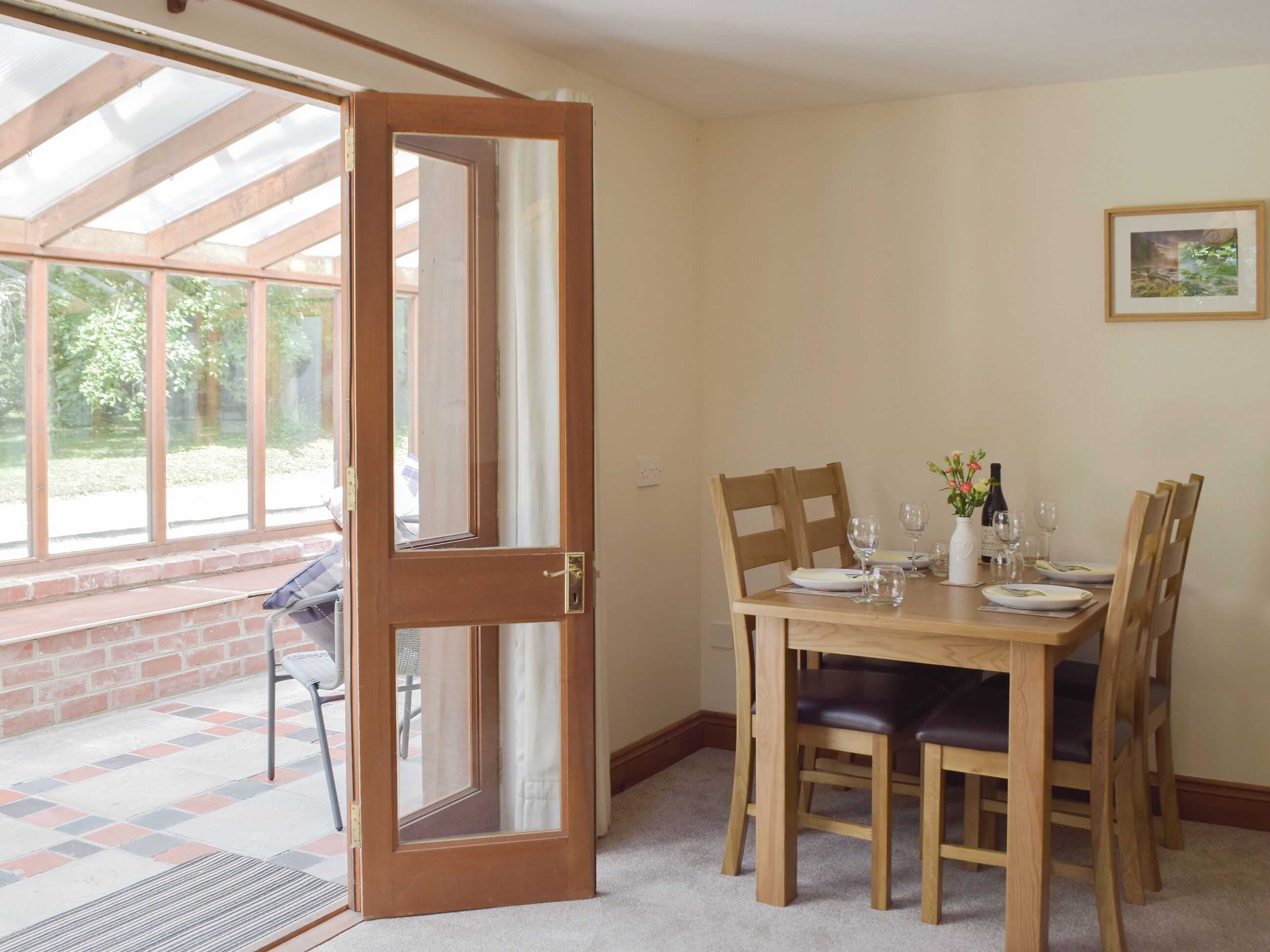 Un coin repas avec une table en bois dressée pour quatre personnes, une porte donnant sur un patio et un tableau encadré accroché au mur.