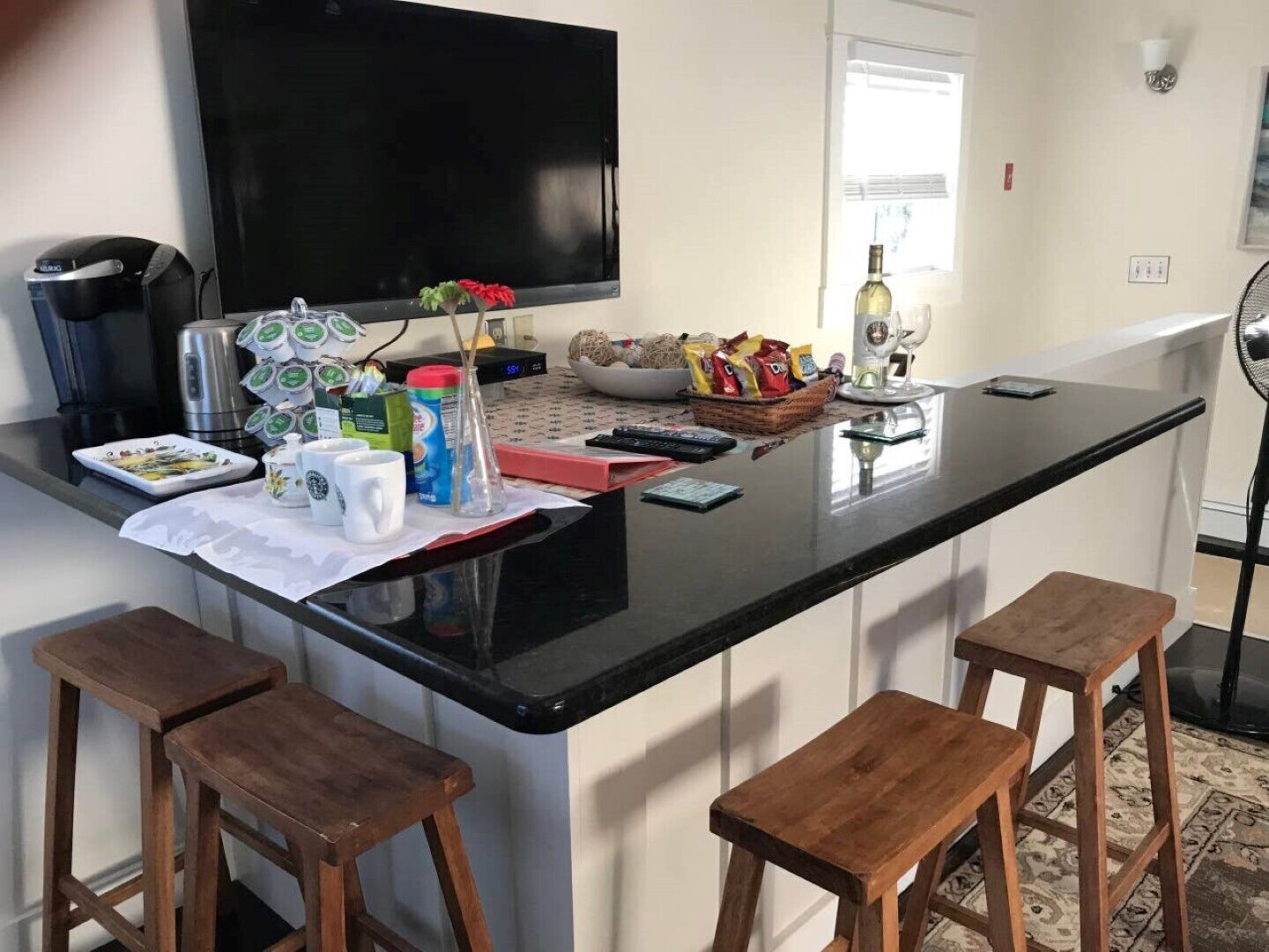 A kitchen with a black countertop, four wooden stools, a coffee maker, a TV, and various kitchen items.