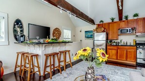 Une cuisine moderne avec des meubles en bois, un réfrigérateur en acier inoxydable et un téléviseur à écran plat. On y trouve deux tabourets en bois et un vase rempli de tournesols, posé sur le comptoir.