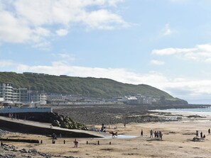 A beach with a rocky shoreline, a promenade with buildings, and a hill with greenery in the background.