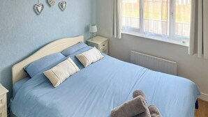 Una cama bien hecha con ropa de cama azul y dos almohadas, una mesita de noche con una lámpara y una ventana con cortinas.