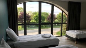 Een hotelkamer met twee bedden, een groot rondboogvenster en uitzicht op groen en een weg.