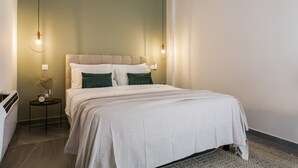 Una cama bien hecha con un cabecero color beige, dos mesitas de noche con lámparas, y un jarrón con flores secas sobre una de ellas.