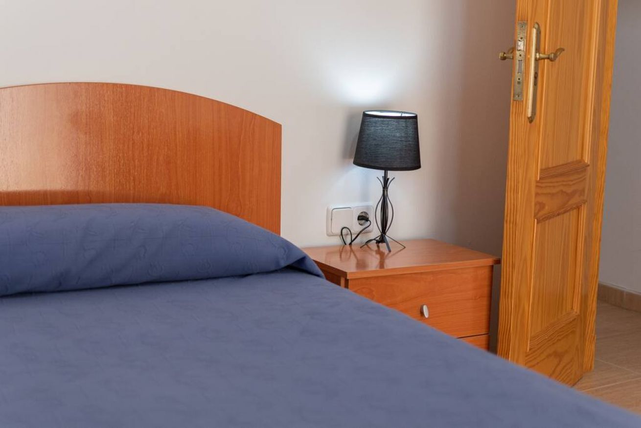 A neatly made bed with a blue bedspread, a wooden nightstand with a lamp, and a wooden door.