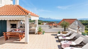 Una terraza con mesa y sillas de madera, un edificio de piedra con techo de tejas y vistas al mar y a las montañas.