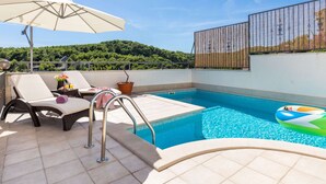 Una piscina en la azotea con sillas de descanso, una sombrilla y vistas a una colina boscosa.