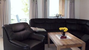 A living room with a black leather sofa, a wooden coffee table with a vase of yellow flowers, and a black armchair. - Bungalow in Boergerende-rethwisch mit Sauna (Boergerende-Rethwisch)