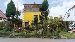 Un edificio amarillo con balcón, rodeado de diversas plantas y árboles.